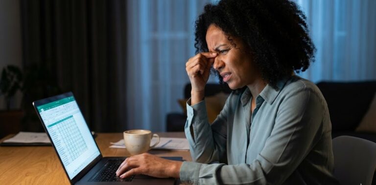 A stressed Afro-Caribbean woman sitting at a desk with a laptop and coffee mug, rubbing her temple to relieve eye strain and eyelid twitching symptoms.